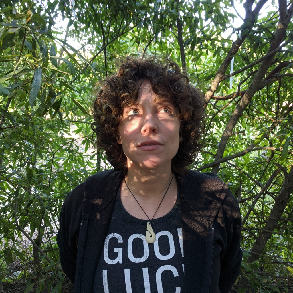A woman with dark curly hair stands under a live willow hut. She is dressed in a black hooded sweatshirt and wears a traditional Maori carving on her necklace. Her face is dappled by sunlight and her eyes look off to the right-hand corner of the frame.